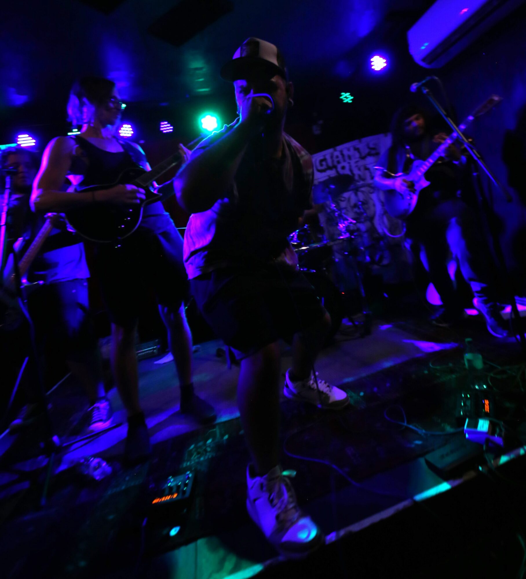 Band performing on stage with colorful lighting, singer in center holding microphone, other musicians playing instruments, dark background.