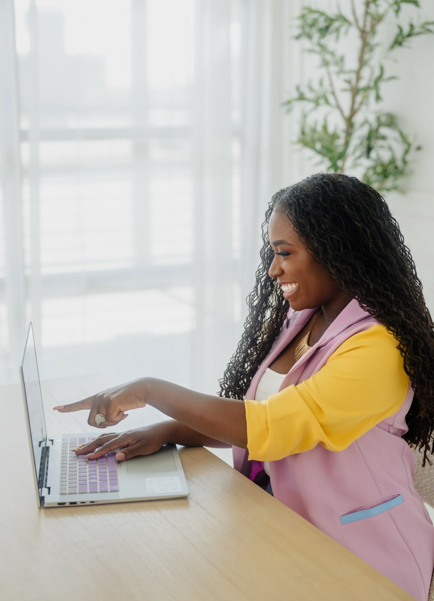 Smiling woman with curly hair using a laptop at a desk, with a plant and window in background.