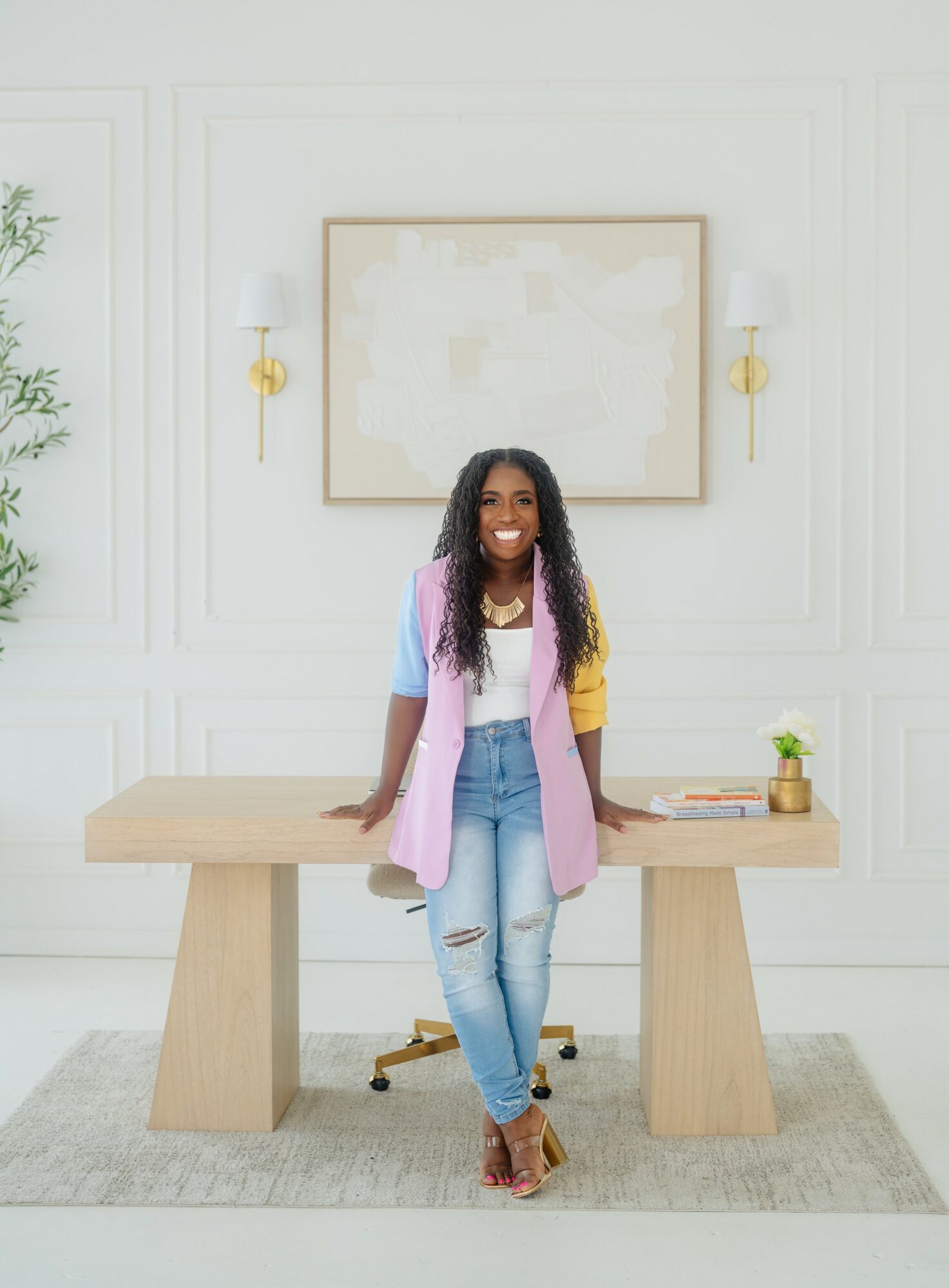 Woman with long curly hair smiling, standing behind a wooden desk in a bright room with white walls and wall sconces.