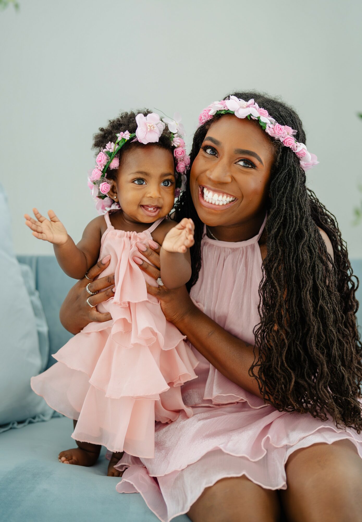 Smiling woman and young girl wearing pink dresses and floral headbands, sitting on a light blue couch, holding each other.