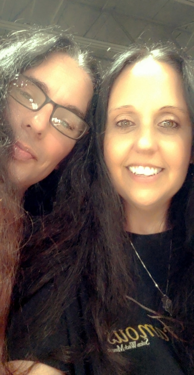 Two women smiling, one wearing glasses, in an indoor setting with a dark background.