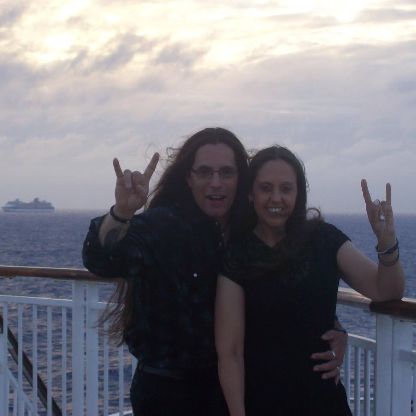 Two people on a boat deck making rock hand gestures, with water and sky in the background.