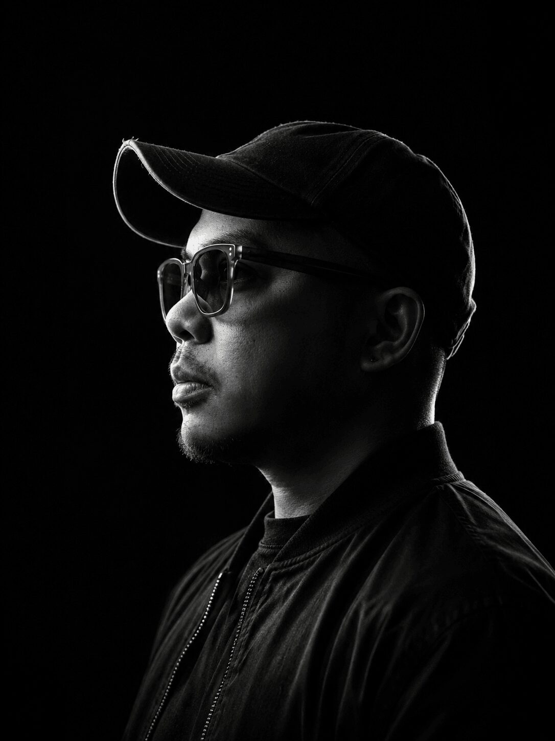 Man wearing sunglasses and a cap, facing left, in black and white with a dark background.