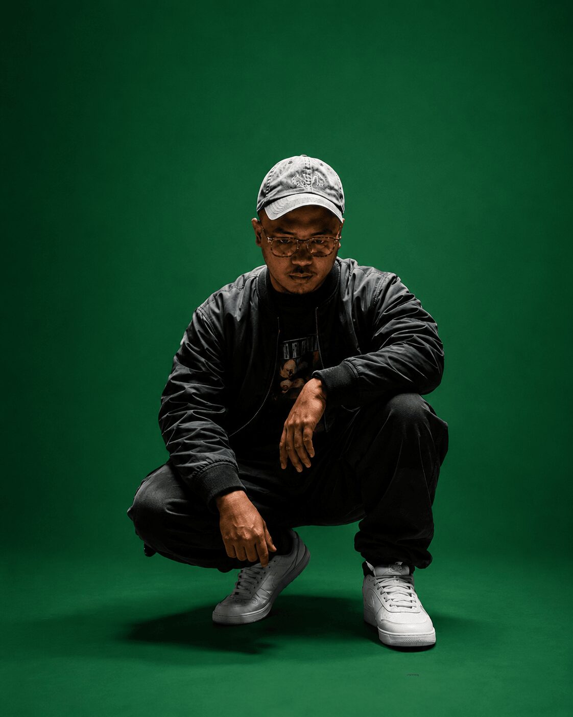 Person crouching in front of a green background, wearing a cap, glasses, black jacket, and white sneakers.