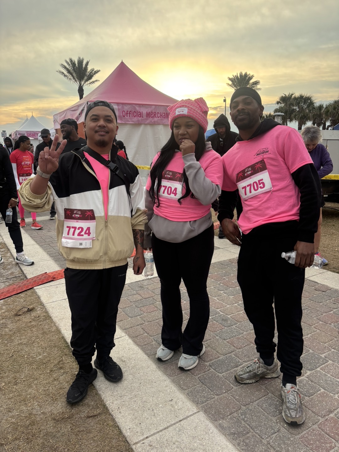 Three people standing outdoors in front of a pink tent, wearing pink shirts and race bibs, smiling at the camera.