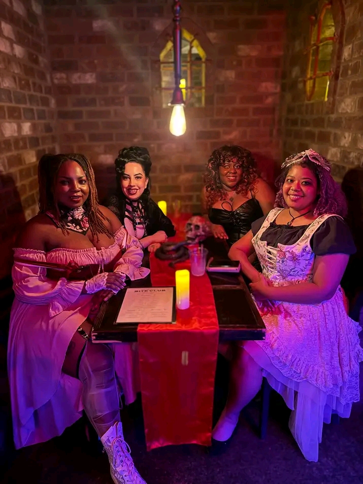 Four women in costumes sit at a table with a red tablecloth, candle, and menu in a brick-walled room with hanging light fixtures.