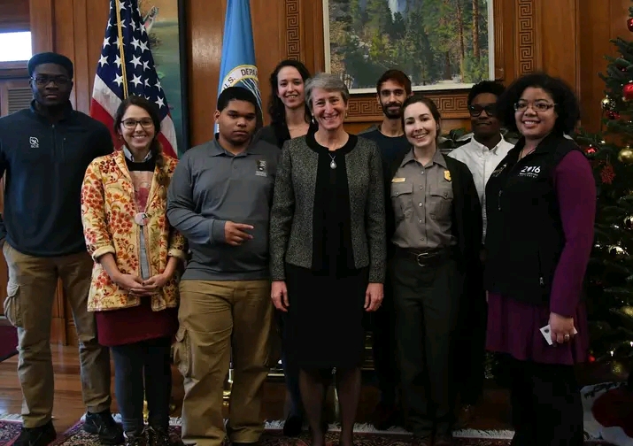 Group of nine diverse people standing indoors, smiling, with flags and a Christmas tree in the background.