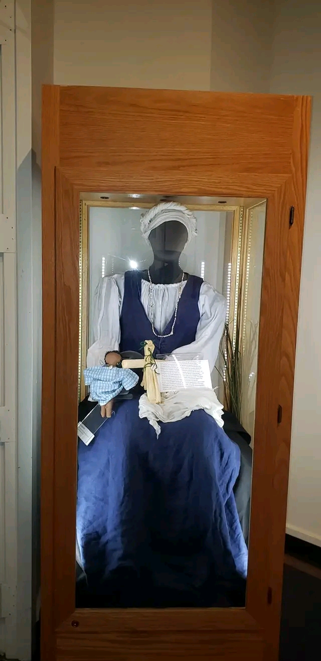 Mannequin dressed in a blue and white outfit inside a wooden display case, viewed from the side.