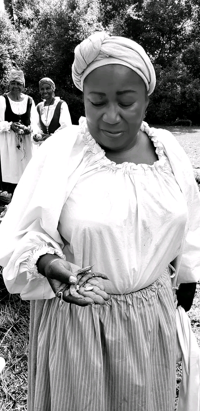 Woman with headwrap and dress holding hands, two women in background outdoors, trees, black and white image.
