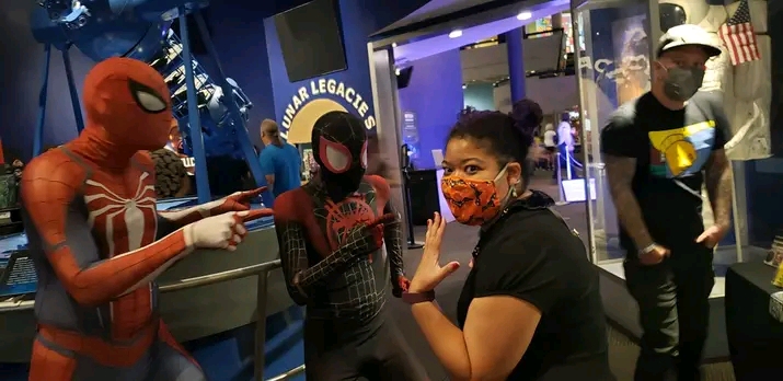 Four people, including two in Spider-Man costumes, pose with a woman at an indoor event. One person wears a face mask.