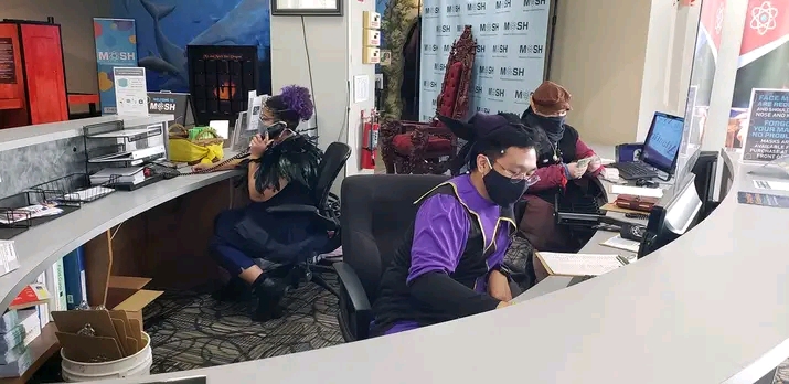 People working at desks with computers and documents in an office environment, some wearing masks.