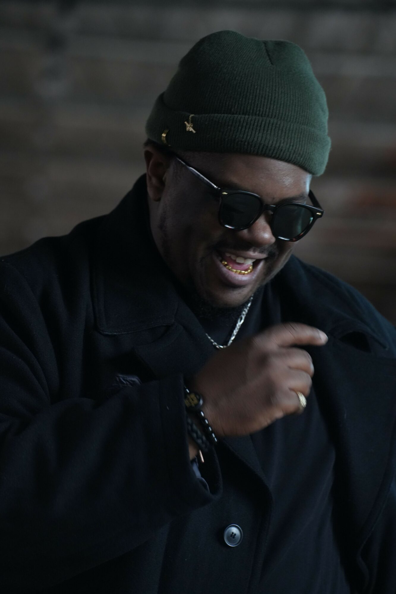Person wearing sunglasses and a green beanie smiling, with a dark jacket, outdoors.