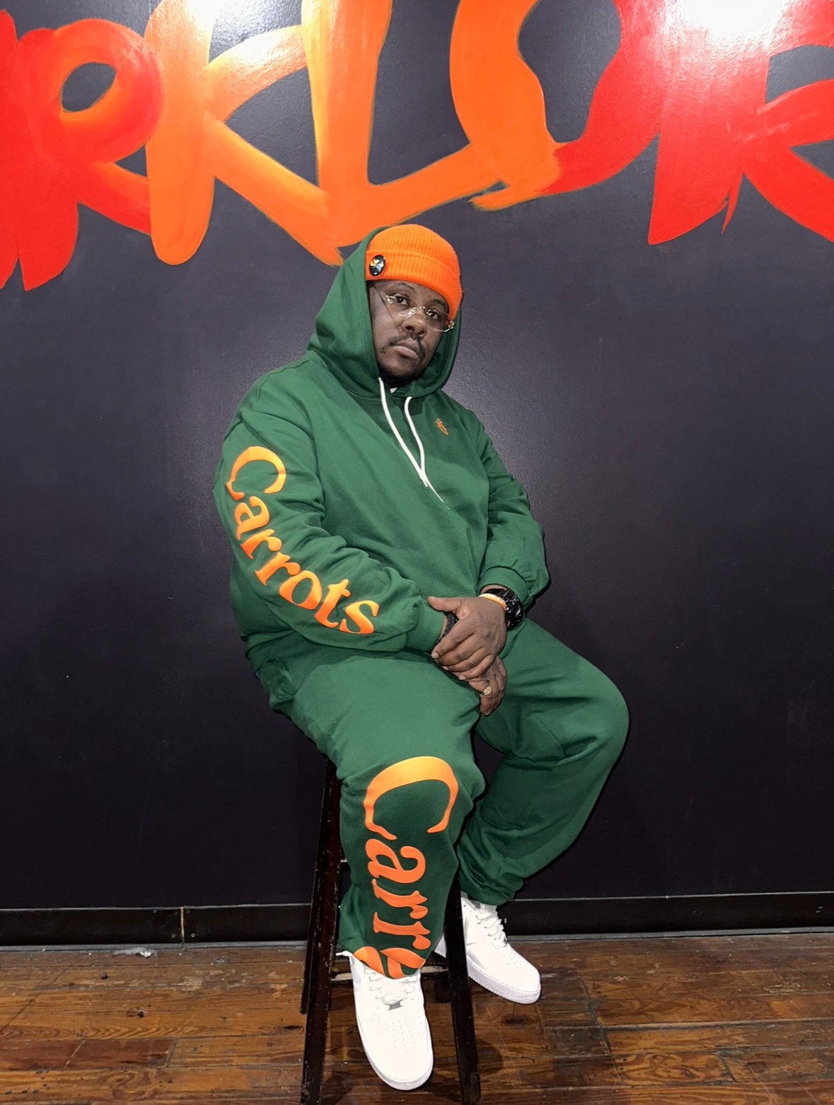 Man in green hoodie and pants with orange and yellow text, sitting on a stool against a black wall with red and orange graffiti