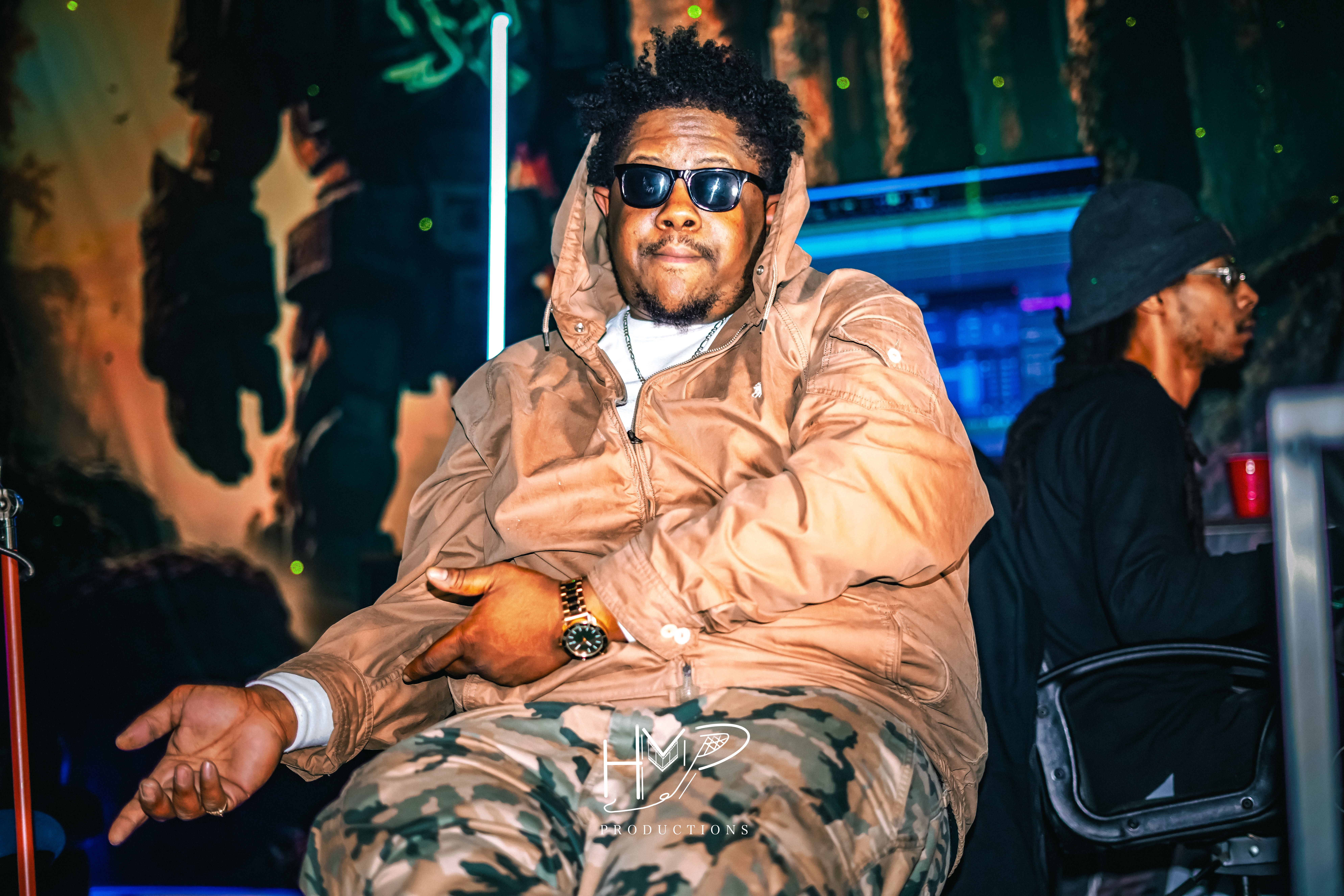 Man wearing sunglasses and a beige jacket sitting with a relaxed pose, background with colorful lights and trees.