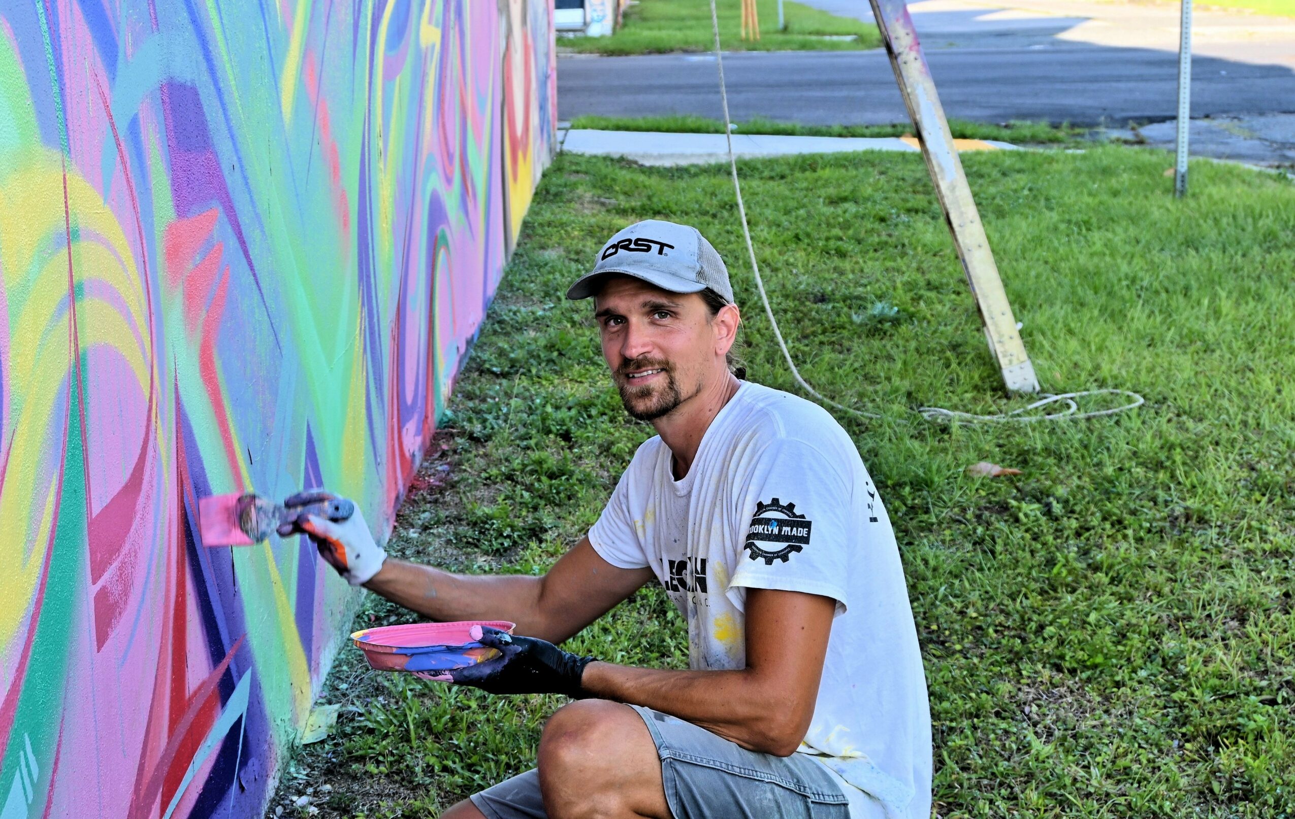 Man painting a colorful mural on a wall outdoors, sitting on grass with a paint palette and brush.