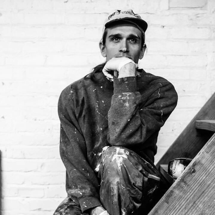Black and white photo of a man sitting on stairs, resting his chin on his hand, wearing a cap and paint-splattered clothes.