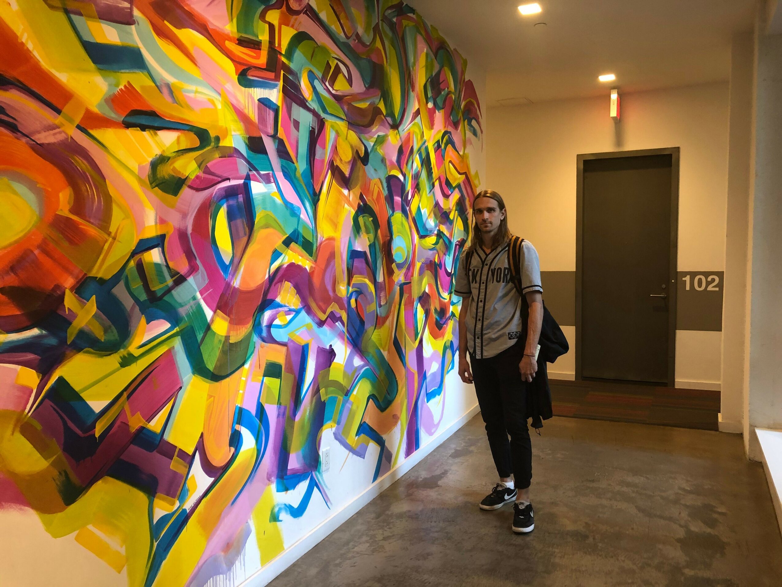 Person standing next to colorful abstract mural on wall, with a window and door in background.