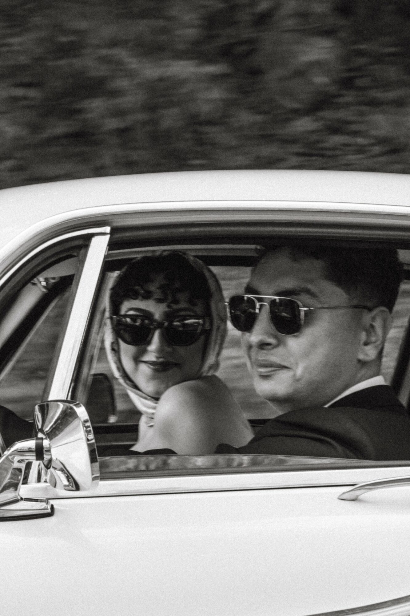Two people wearing sunglasses sit in a car, smiling and looking out the window.