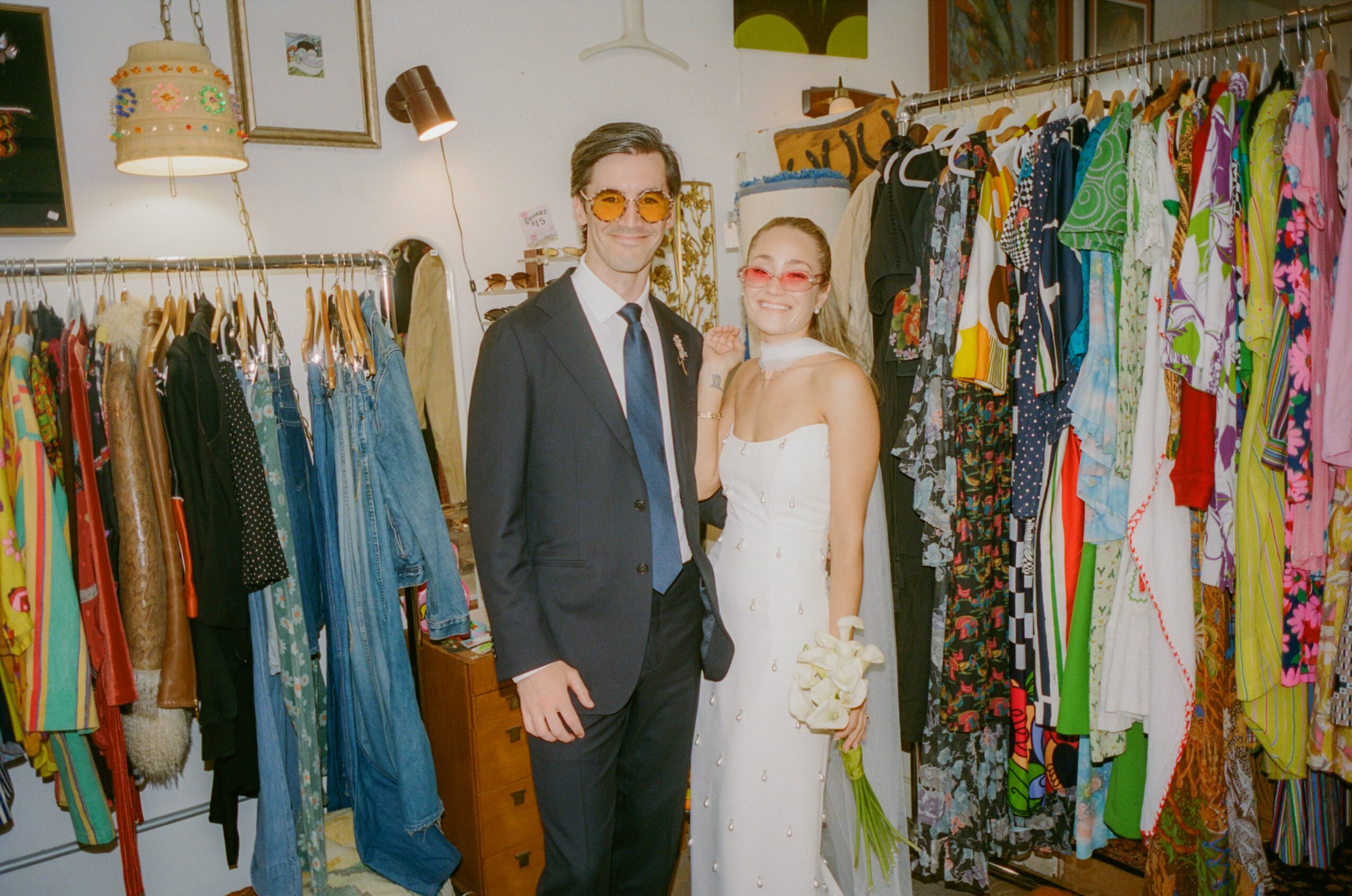 A man in a suit and a woman in a white dress stand together in a clothing store, surrounded by racks of colorful clothes.