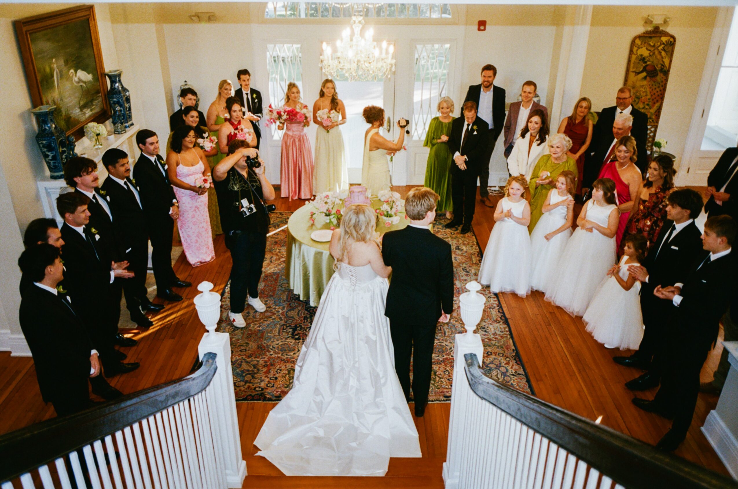 Wedding ceremony with bride and groom at center, surrounded by guests in formal attire, in a decorated room with chandelier.