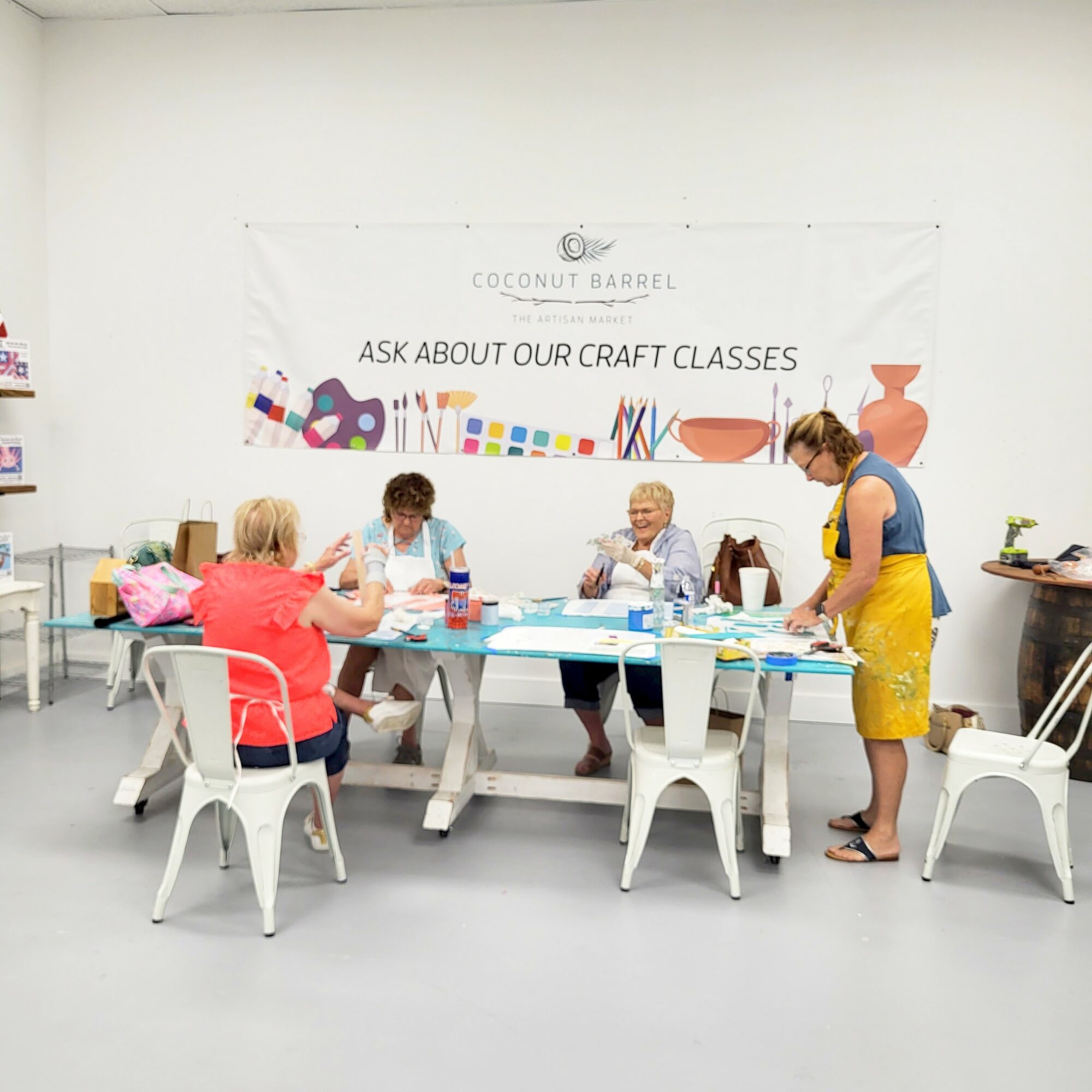 Four people sit around a table with craft supplies, one person stands, in a craft class room with a banner. 20 words.