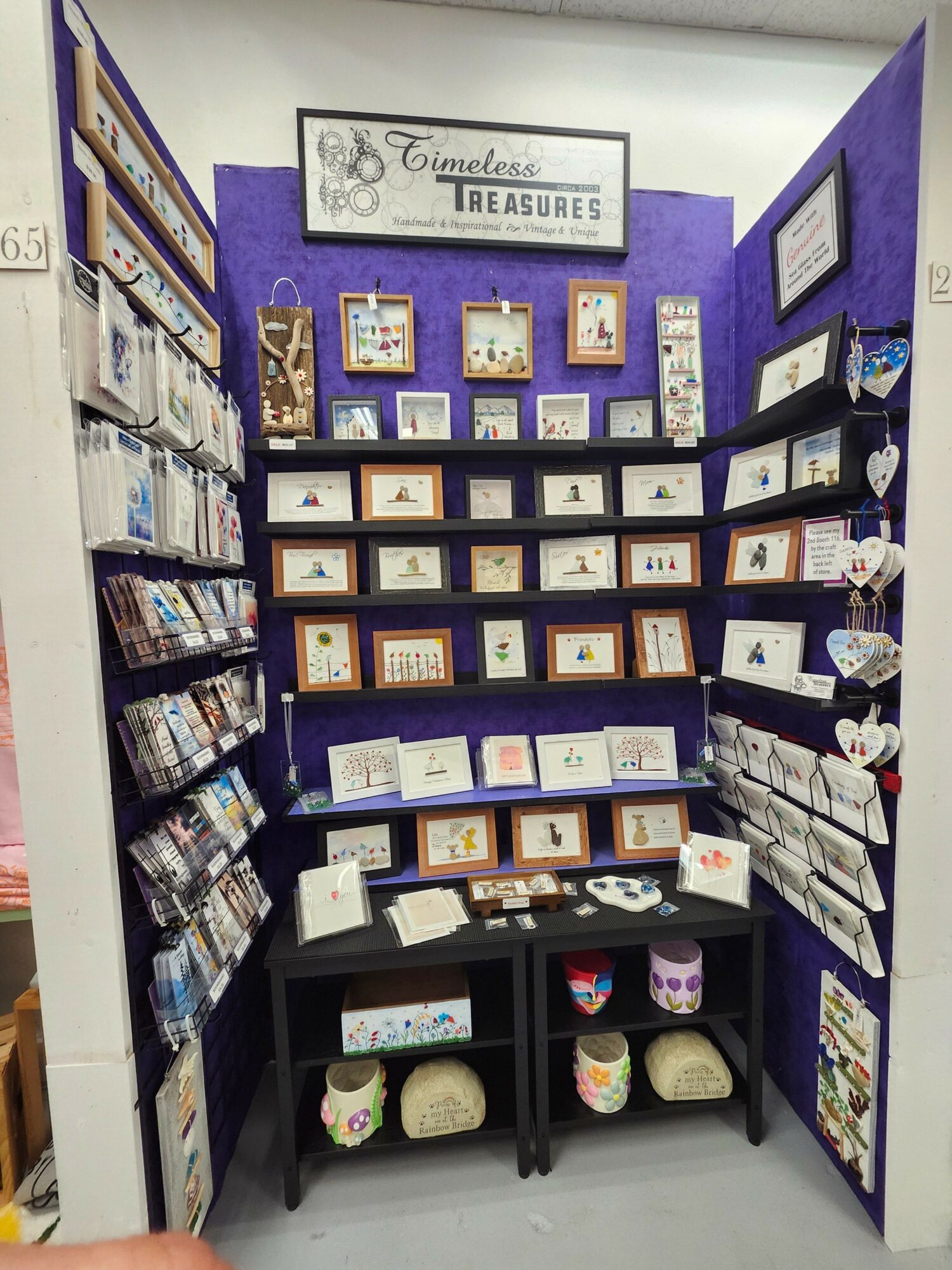 Display of framed artwork, candles, and decorative items on purple shelves with a sign reading 'The Little Things.'