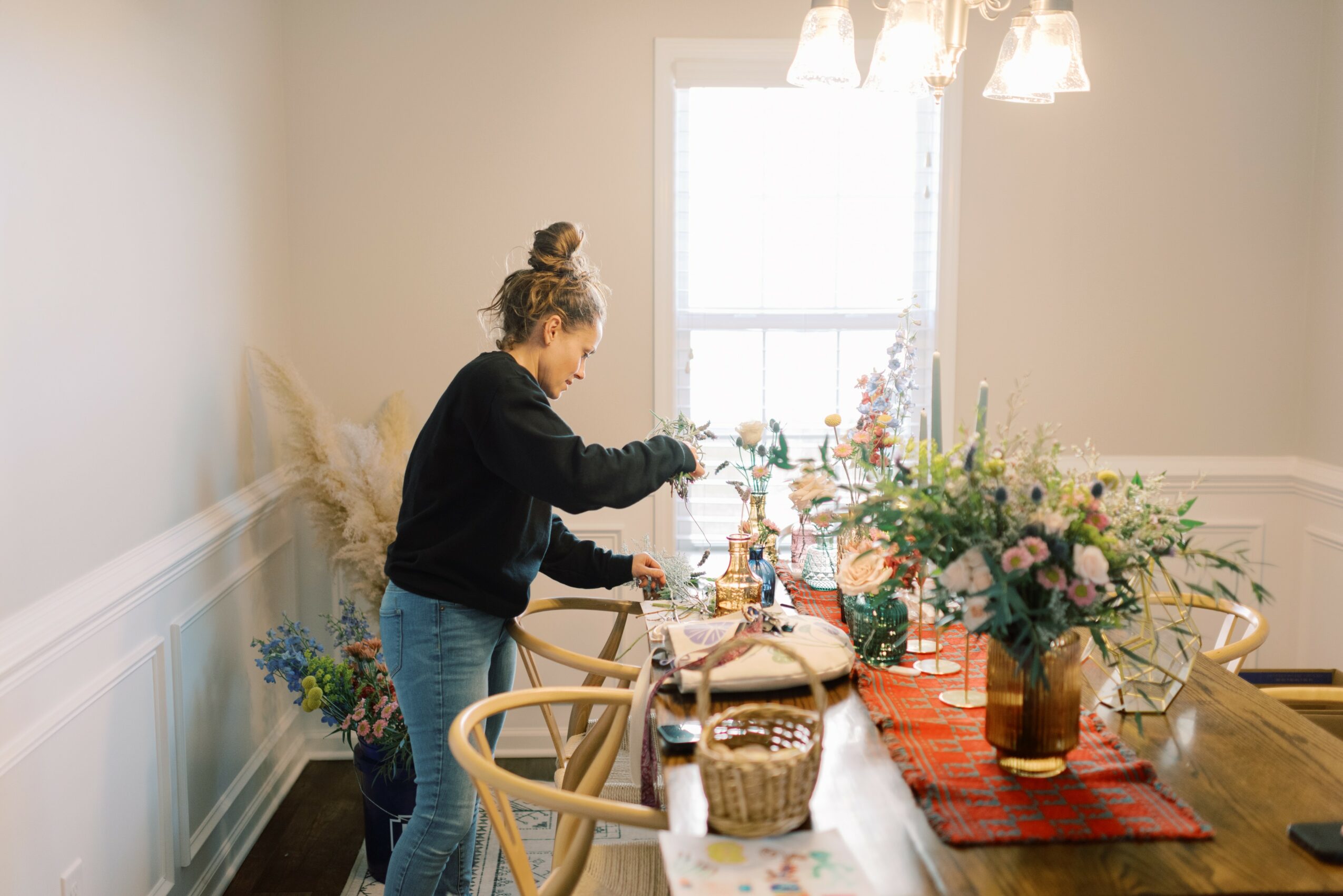 Person arranging flowers on a dining table with a window and chandelier in the background.