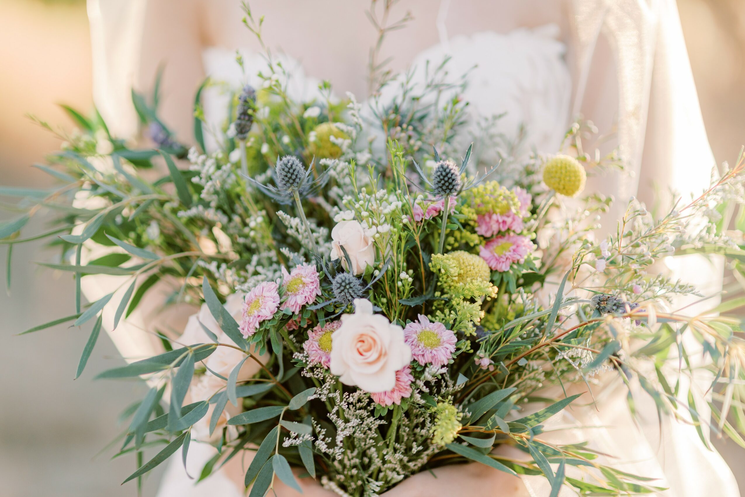 Person holding a large bouquet of mixed flowers with greenery, pink, white, and purple blooms, outdoors in sunlight.