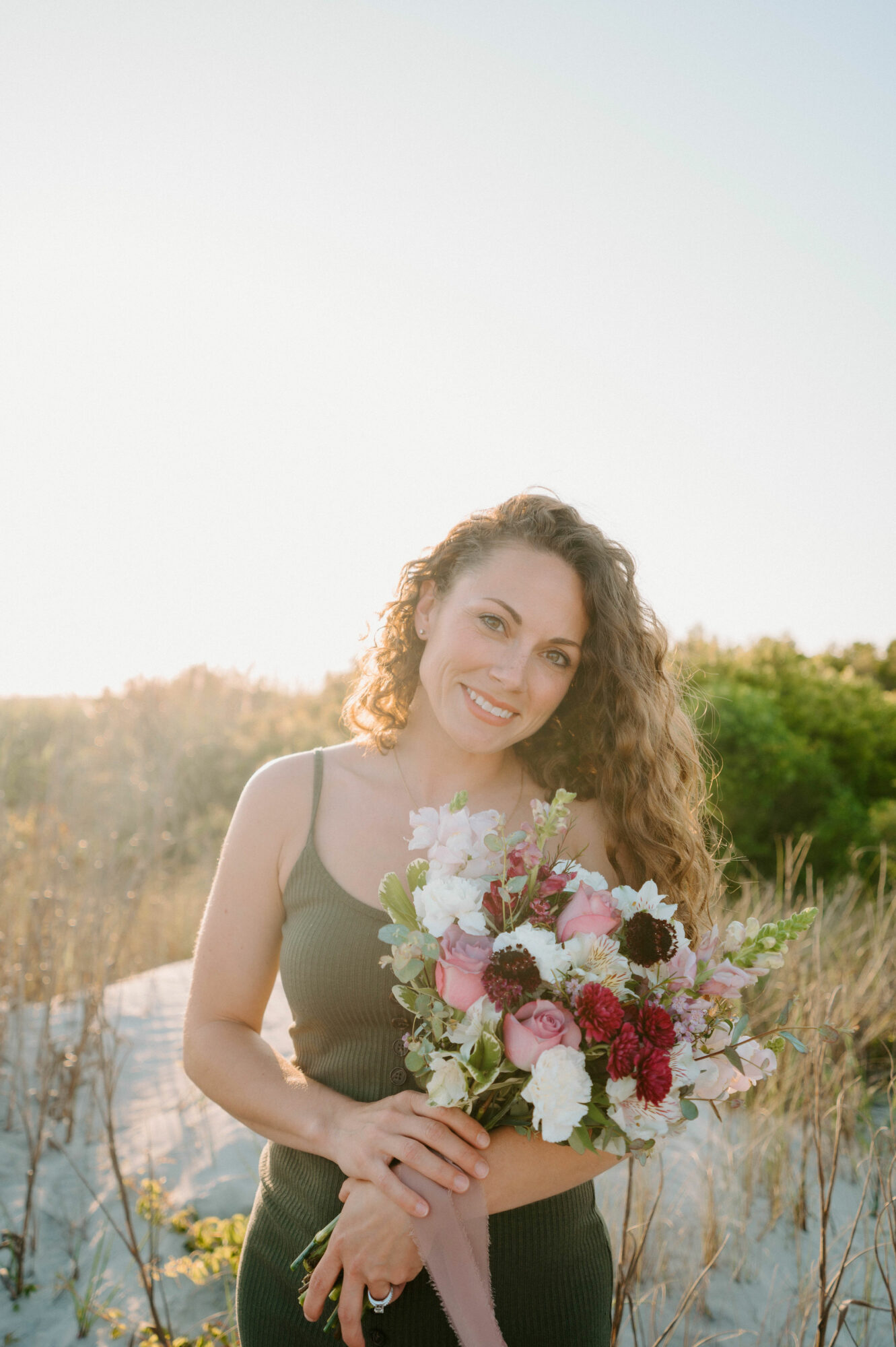 Young woman with long curly hair holding a bouquet of flowers outdoors, smiling, with a natural background.
