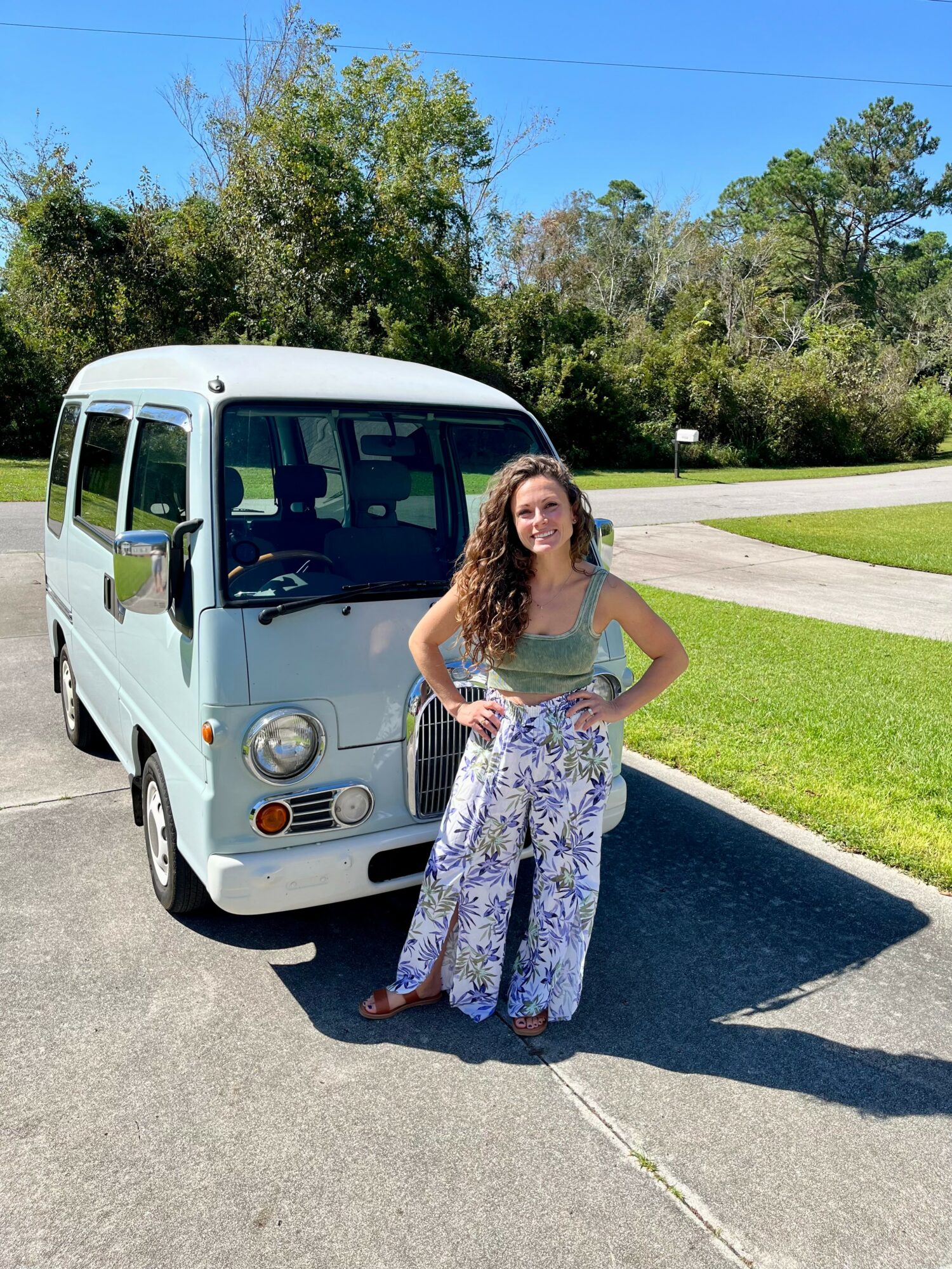 Young woman with long curly hair standing outdoors next to a small white vehicle on a sunny day, trees in background.