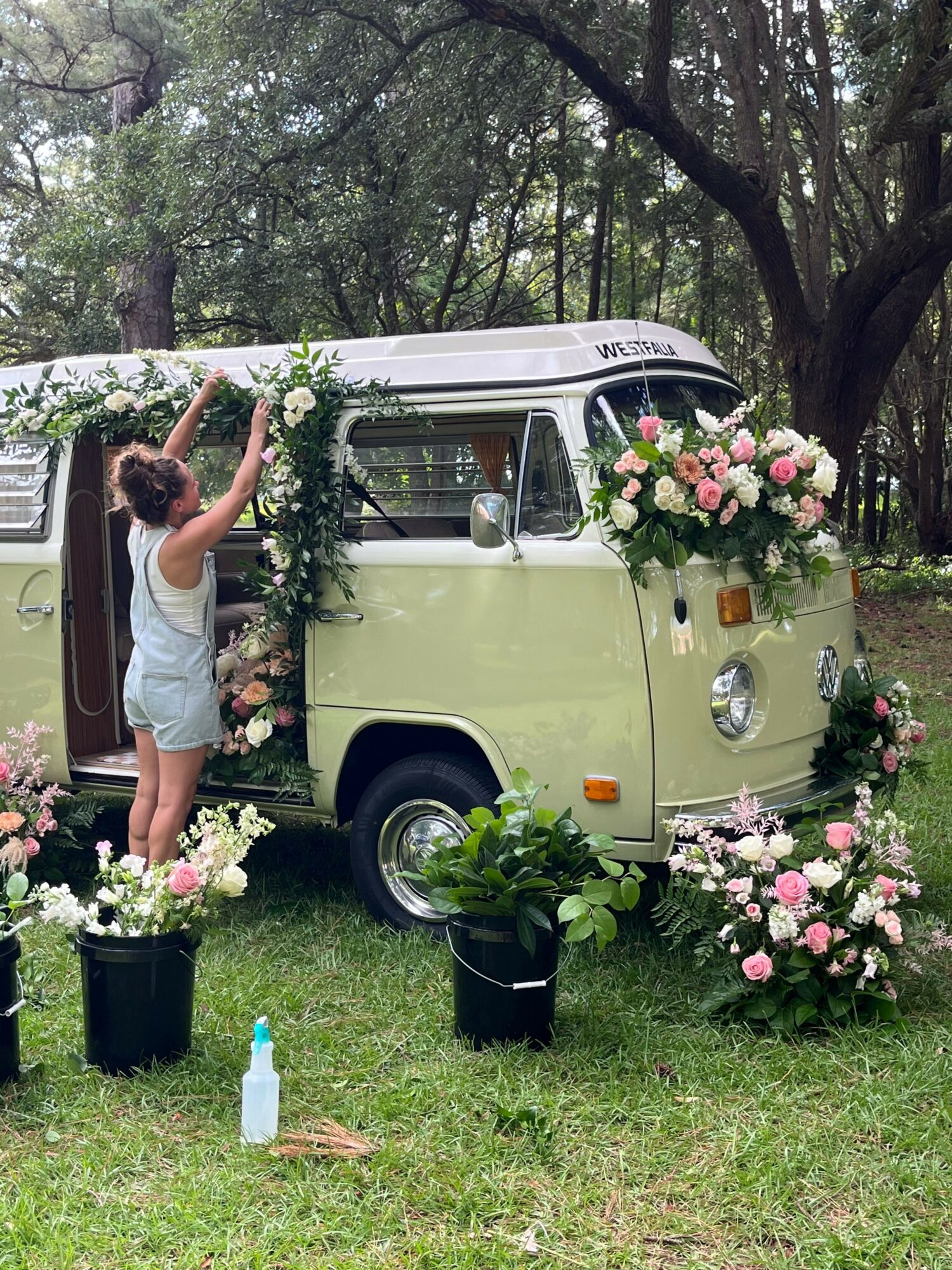 Person arranging flowers on a vintage van decorated with pink and white flowers outdoors.