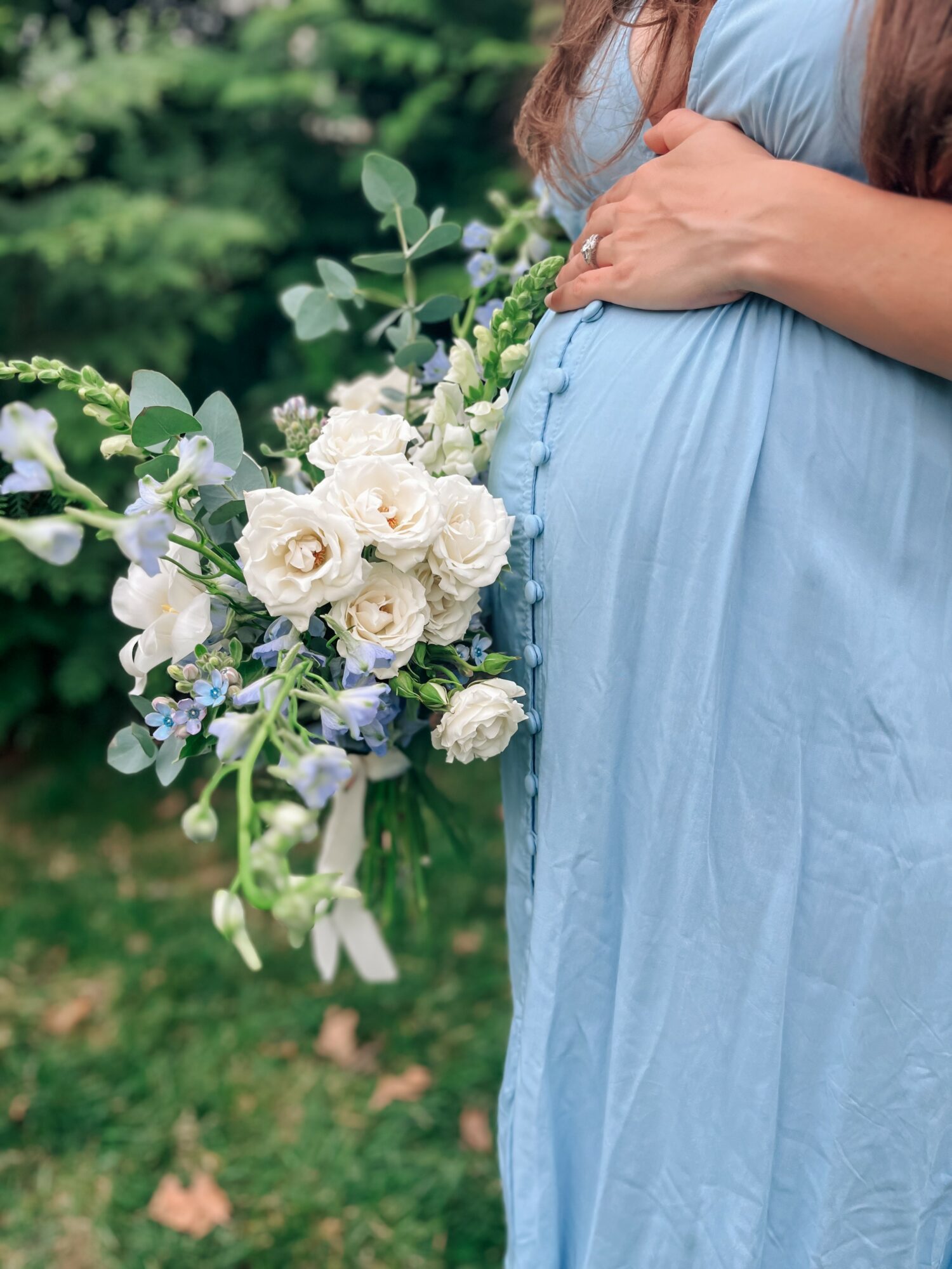 Pregnant woman in a blue dress holding a bouquet of white and purple flowers outdoors.