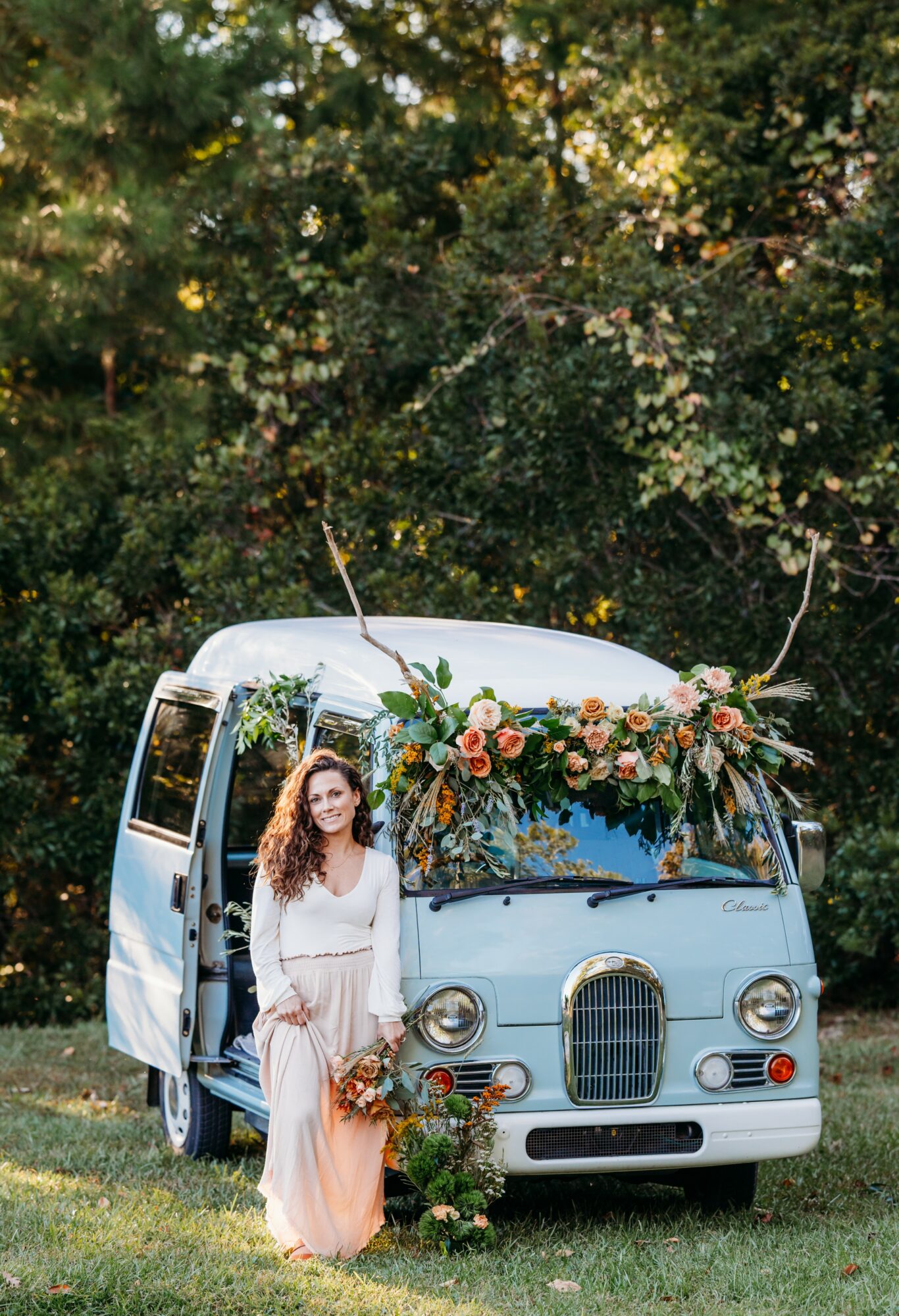 Woman in white dress standing beside a vintage van decorated with flowers outdoors, surrounded by trees and greenery.