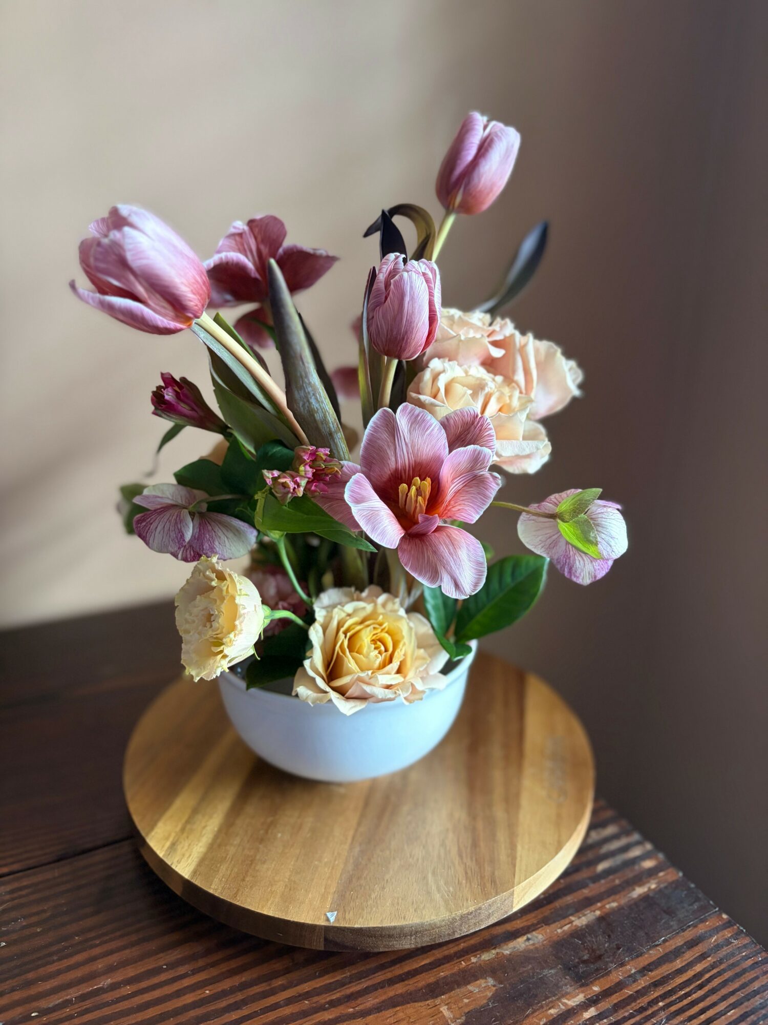 Flower arrangement with pink tulips, roses, and other flowers in a white bowl on a wooden tray.