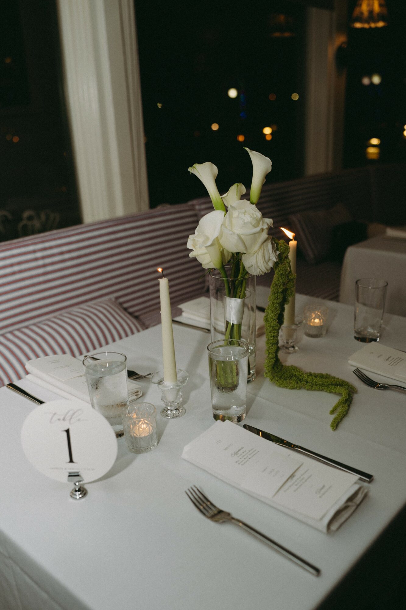 Table setting with a floral centerpiece, candles, glasses, and a menu, near a window with city lights outside.