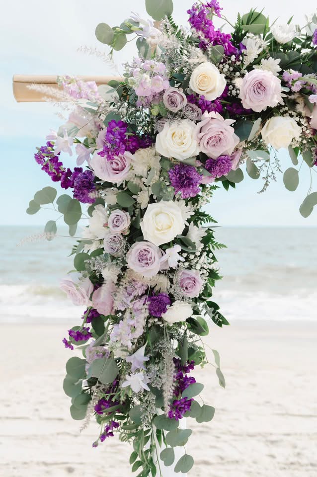 Vertical floral arrangement with white and purple flowers and green foliage, set against a beach background.