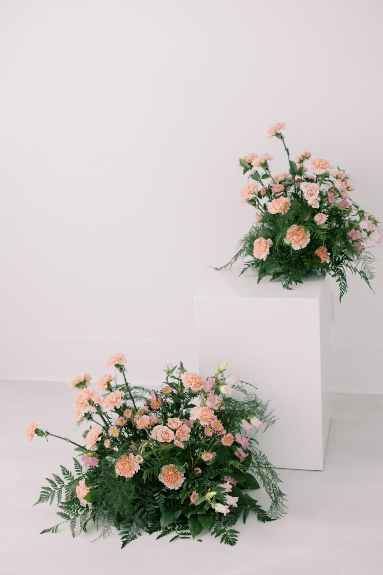 Two floral arrangements with pink and peach flowers and green foliage, one on the floor and one on a white pedestal, against a plain white background.
