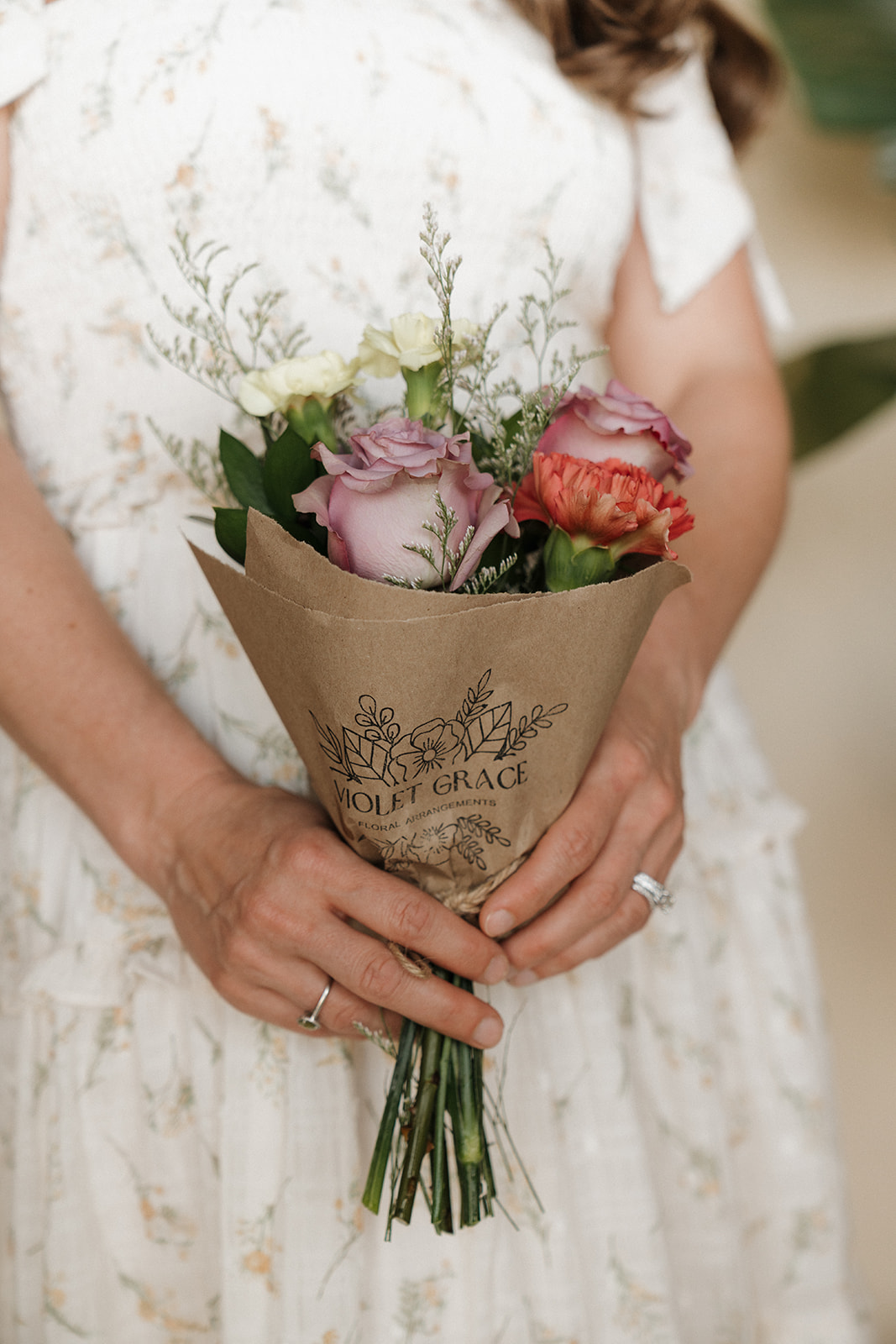 Person holding a bouquet of pink, red, and white flowers wrapped in brown paper, with a white dress and blurred background.