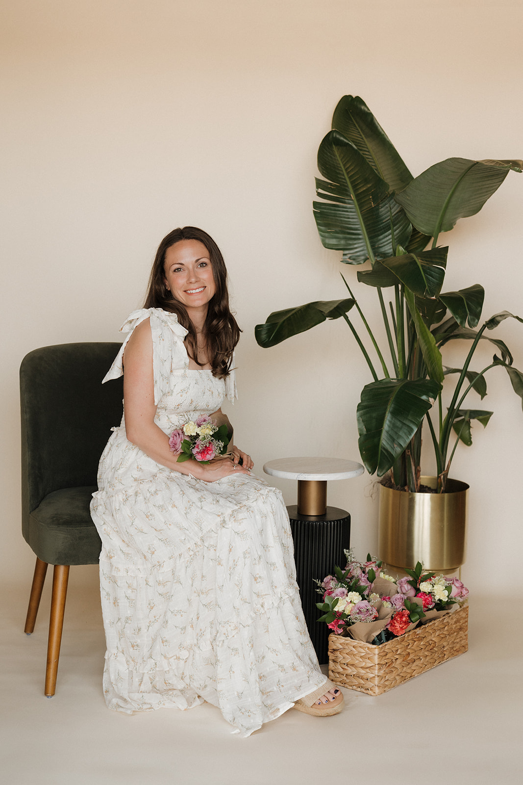 Woman in a white dress sitting on a dark green chair, holding a bouquet, next to large potted plant and flowers.