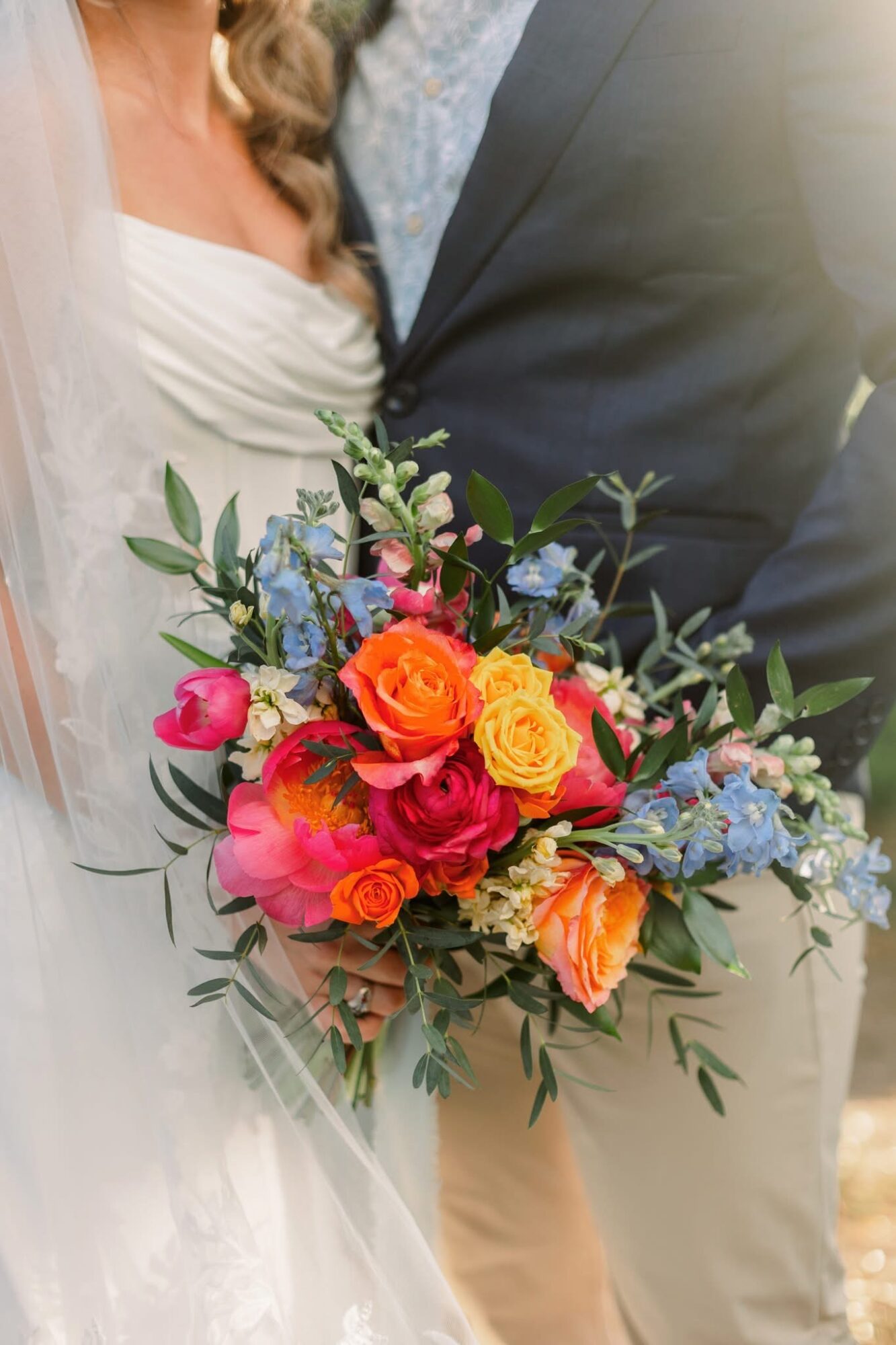 Bride holding a colorful bouquet of roses and greenery, with part of her white dress visible, and a man in a suit nearby.