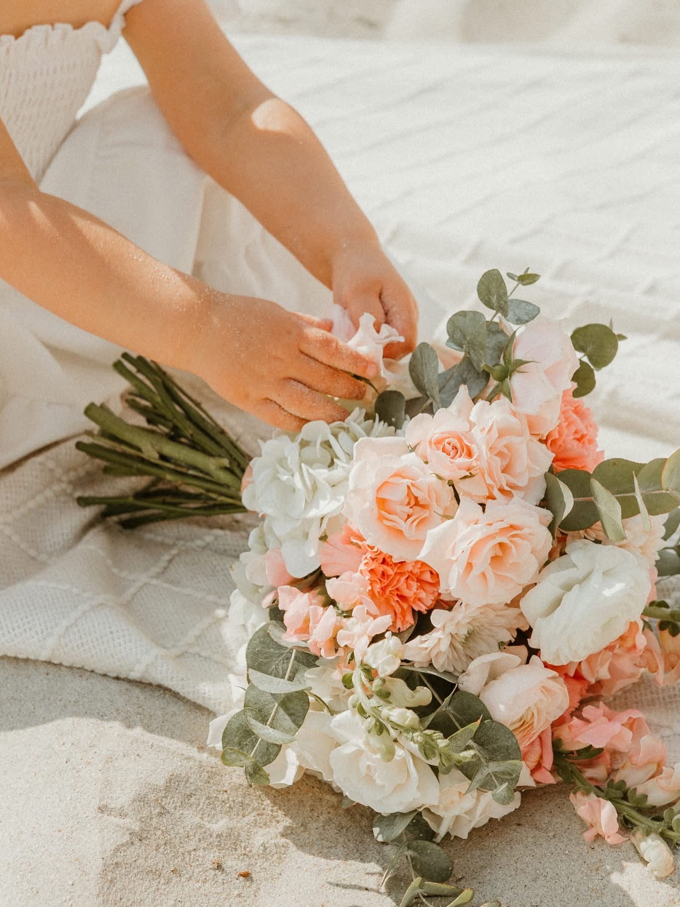 Person arranging a pastel-colored bouquet of roses, peonies, and greenery on a white surface outdoors.