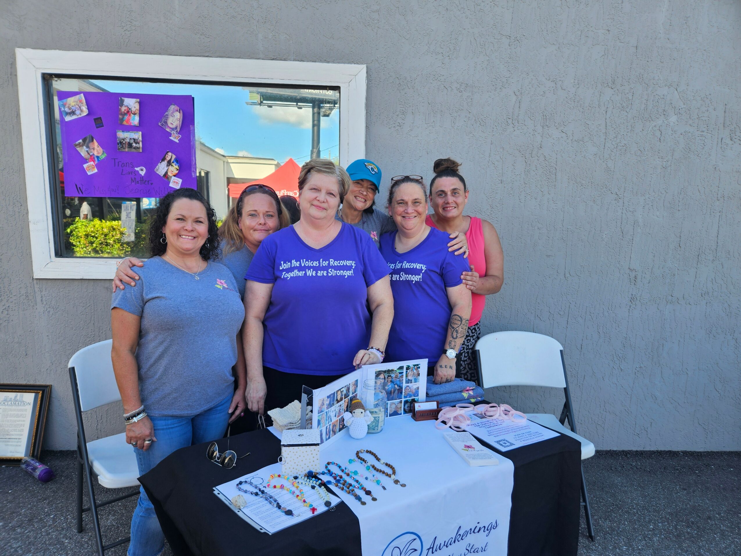 Group of six women standing behind a table with promotional materials outside a building, smiling, with a window reflecting a blue sky.