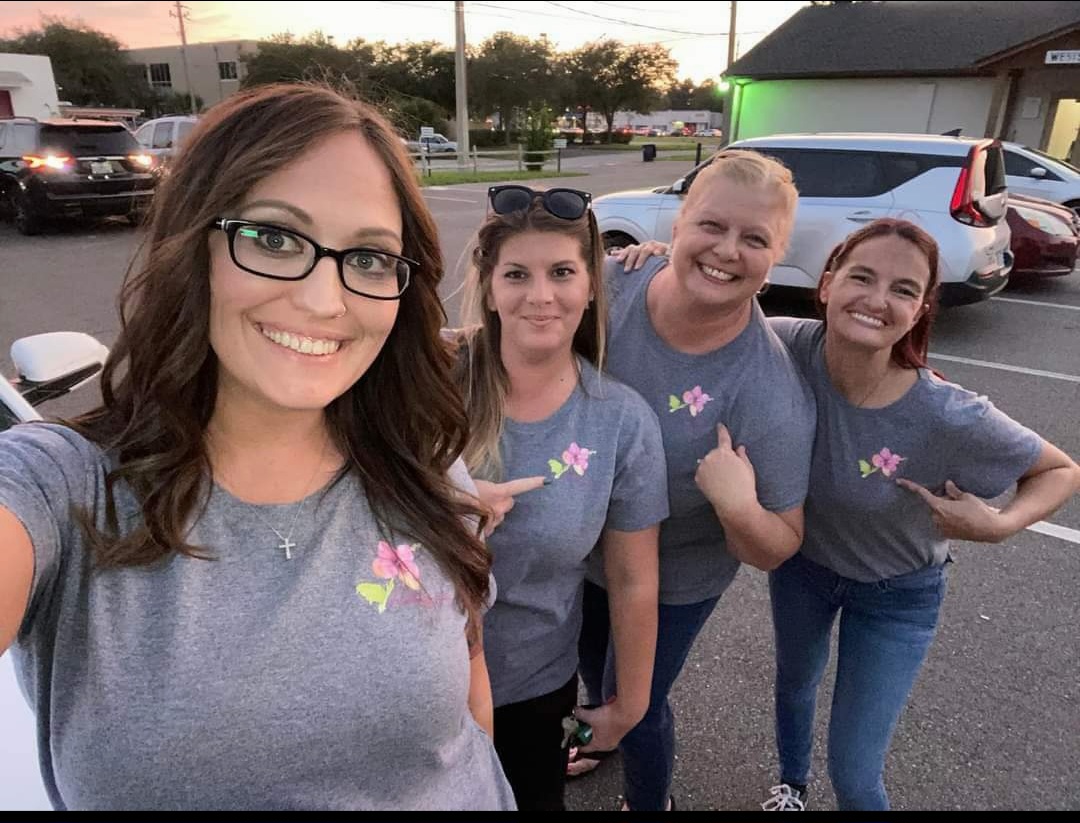 Four women standing outdoors, smiling, wearing matching gray shirts with pink and yellow flower designs, in a parking lot.