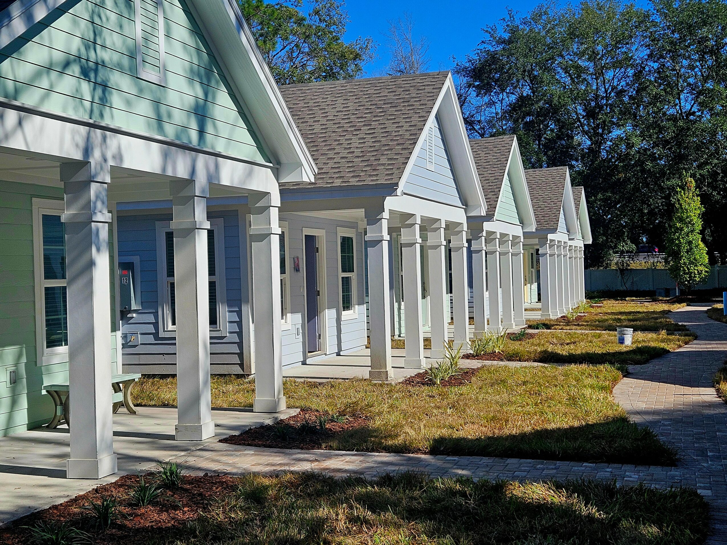 Row of houses with porches and white columns, grassy lawns, and a sidewalk, under a blue sky with trees in the background.