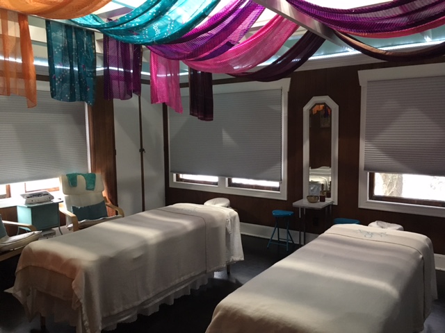 Two massage tables in a room with colorful fabric drapes on the ceiling, windows with blinds, and a mirror on the wall.