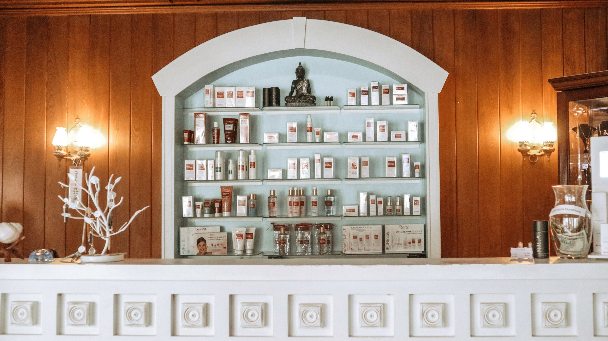 Shelves filled with various bottles and containers behind a white counter, with wooden walls and wall-mounted lights.