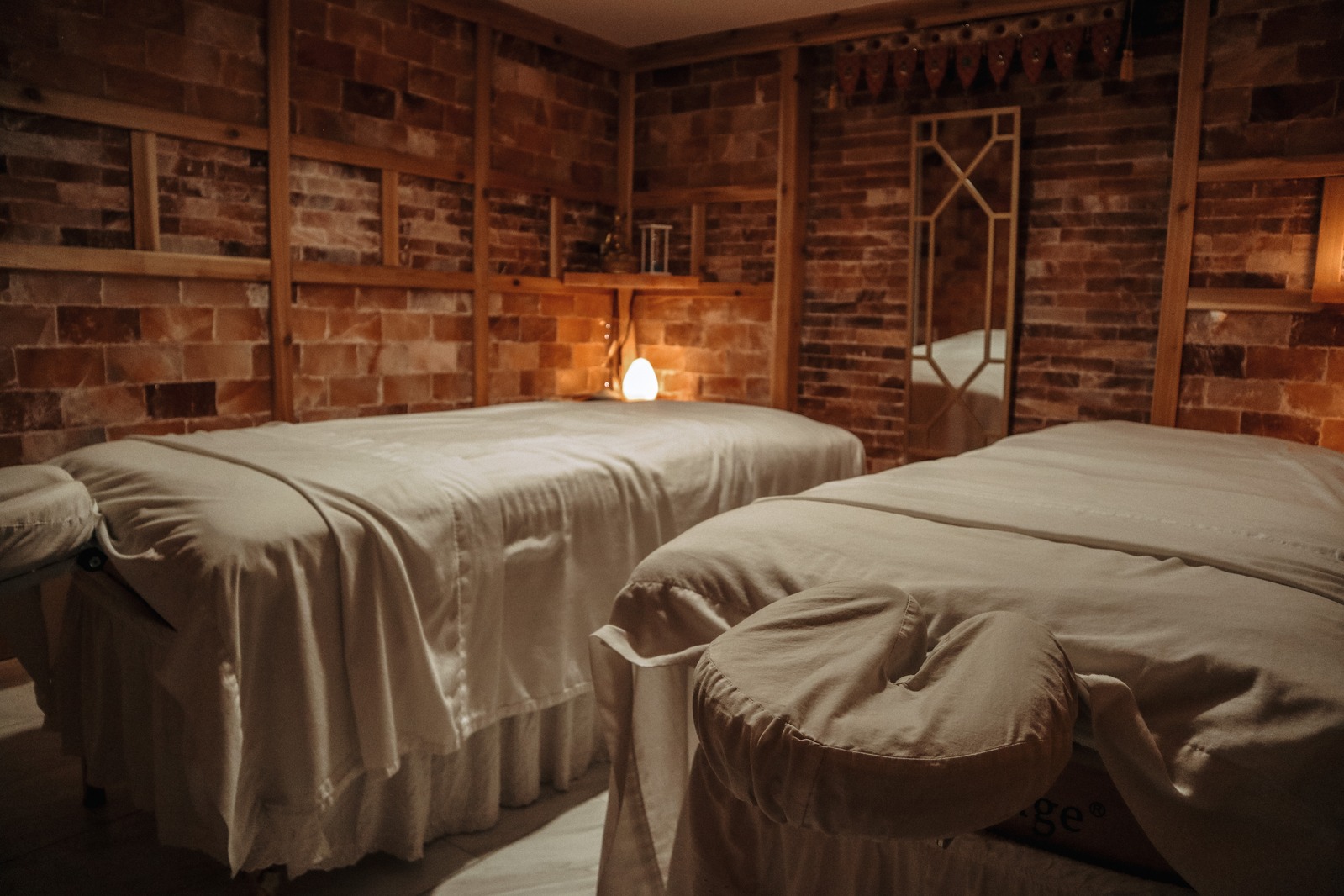 Two massage tables covered with white sheets in a room with brick walls and wooden shelves.