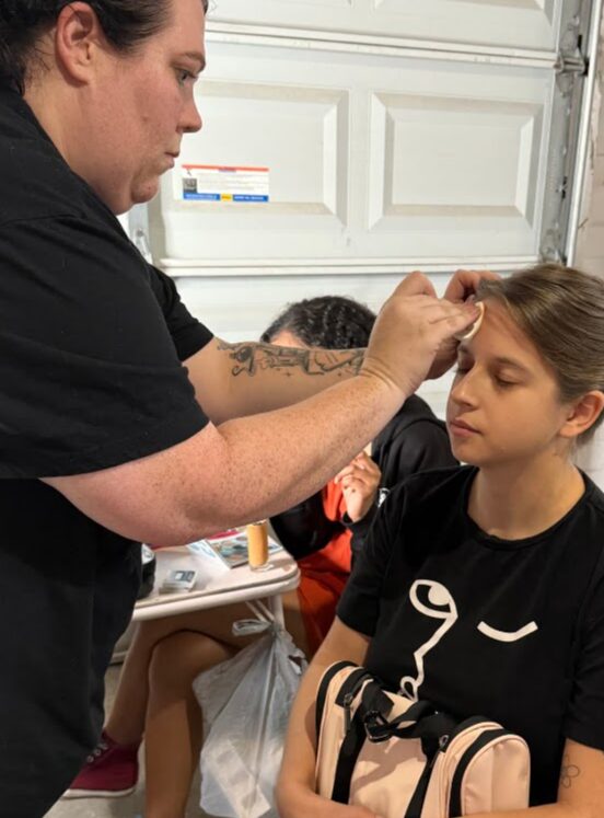 Adult woman applying makeup or face paint to a young girl with closed eyes, in a garage or workshop setting.