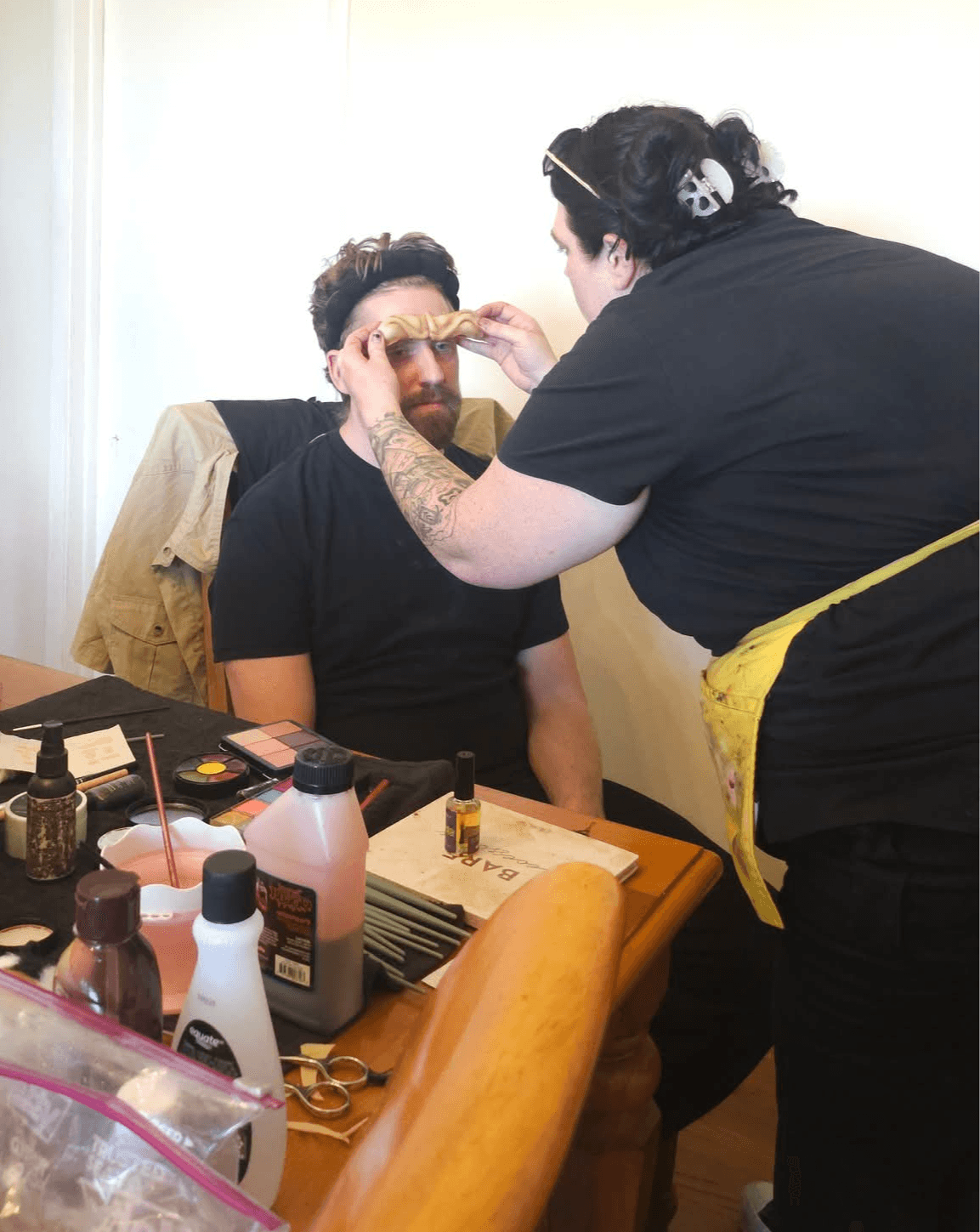 Person sitting with eyes closed as another person applies makeup or face paint to their forehead, surrounded by various bottles and tools on a table.