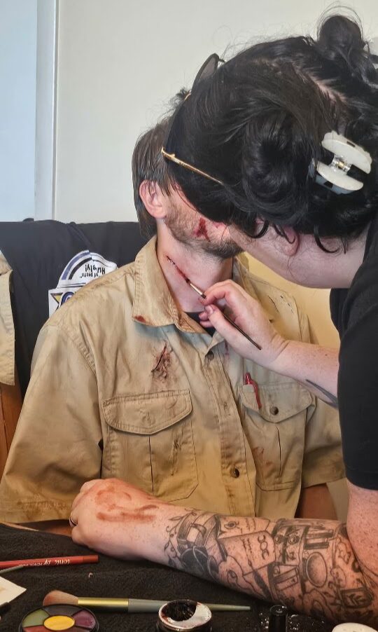 Person with makeup applied to face, seated at table, with another person adjusting makeup, tattooed arm visible.